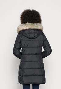 Žena v černém prošívaném kabátu s kapucí lemovanou kožešinou, stojící zády k jednoduchému světlému pozadí.