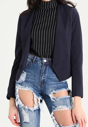 Mujer con un cuello de cisne negro de rayas, blazer azul marino y vaqueros azules de talle alto muy rasgados con grandes desgarros en los muslos.