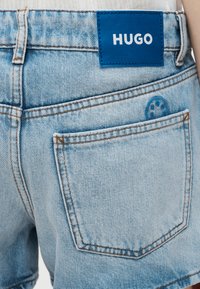 Ανοιχτό μπλε σορτς τζιν με πίσω τσέπη και γαλάζια ορθογώνια ετικέτα HUGO ραμμένη πάνω από τη μέση.