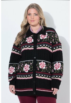 Kvinde iført en sort knapjakke med pink og hvide blomsterprint over en ternet skjorte og bordeauxbukser.