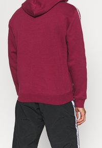 Uomo che indossa un felpa bordeaux con cappuccio abbassato e pantaloni sportivi neri con una striscia bianca laterale, visto da dietro.