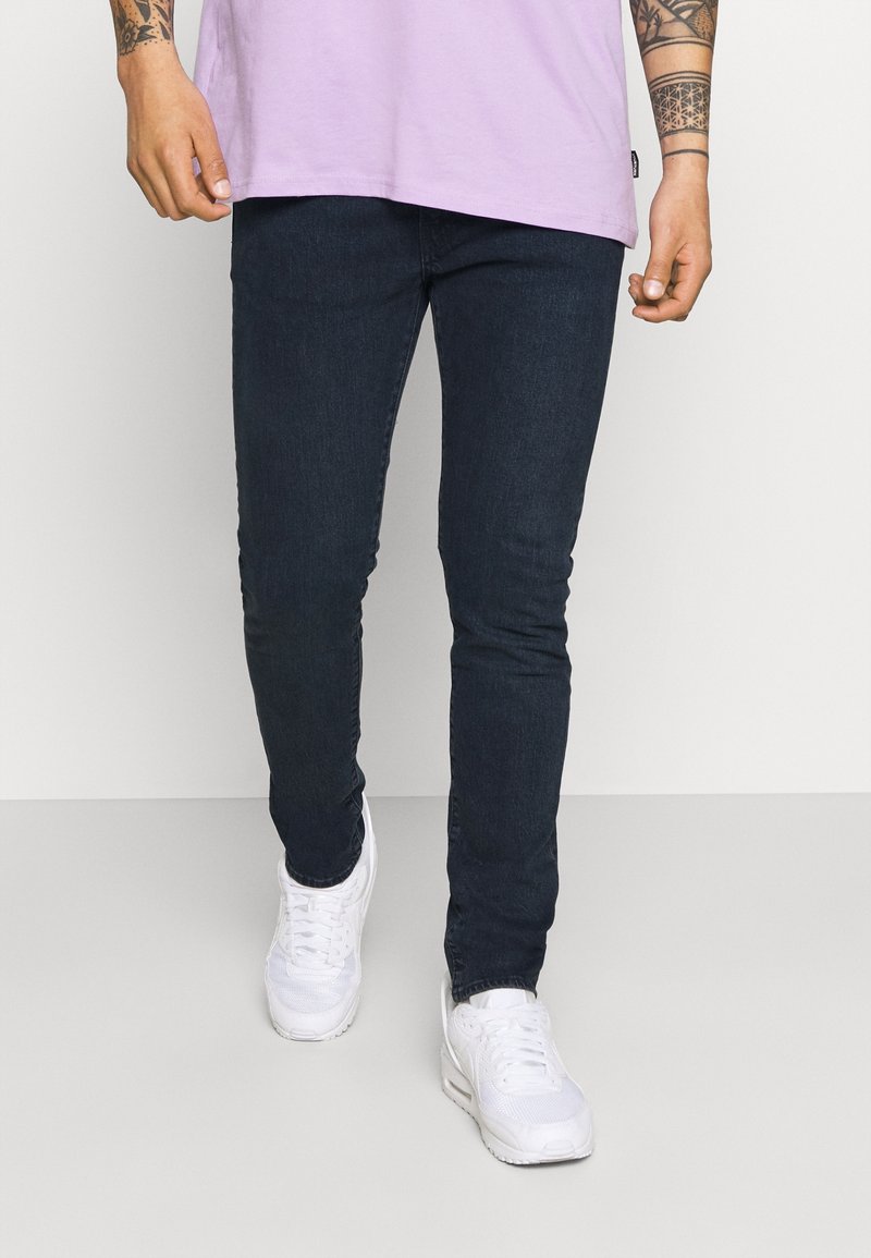 Person som bär mörkblå slim-fit jeans, en ljuslila skjorta, vita sneakers och synliga tatuerade underarmar medan de går framåt.