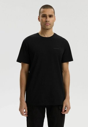 Mężczyzna o krótkich włosach w jednolitej czarnej koszulce i czarnych spodniach, stojący na jednolitym jasnoszarym tle.