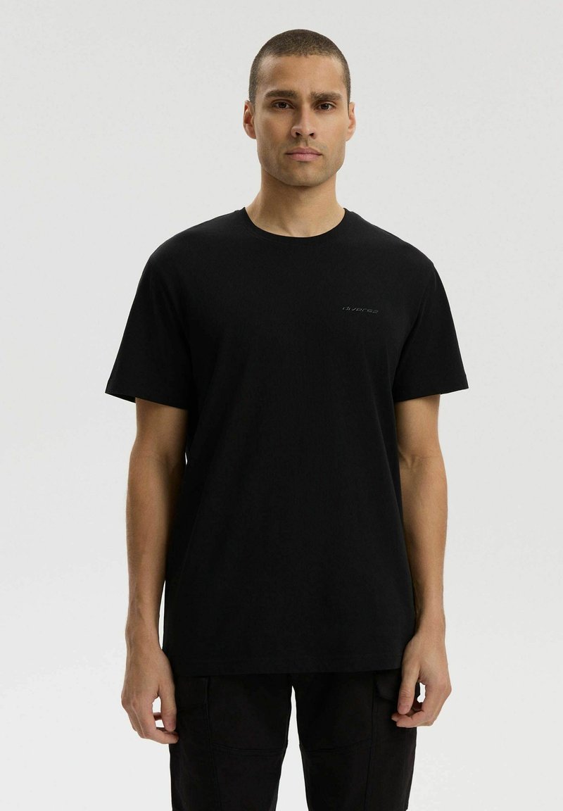 Mężczyzna o krótkich włosach w jednolitej czarnej koszulce i czarnych spodniach, stojący na jednolitym jasnoszarym tle.