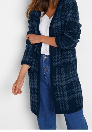 Donna che indossa un cardigan a quadri blu navy sopra una camicia bianca con bottoni e jeans blu a vita alta con chiusura a bottoni.