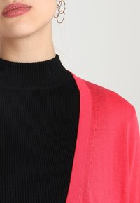 Schwarzer, taillierter Rollkragenpullover unter einer rosa Strickjacke, mit V-Ausschnitt und seitlichen Nähten für zusätzliche Textur.