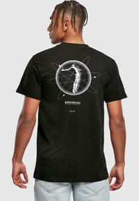 Sort bomulds T-shirt med et grafisk design på ryggen, der viser en astronaut inden i en cirkulær atomillustration med tekst og stregkode.