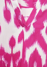 Textil s živým růžovým a bílým ikat vzorem s výstřihem do V. Látka má hladkou texturu a výrazný barevný kontrast.