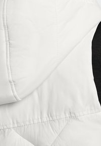 Biała ocieplana kurtka z haftowanym wzorem i kapturem. Miękka, gładka tekstura z kontrastującymi czarnymi akcentami po bokach.