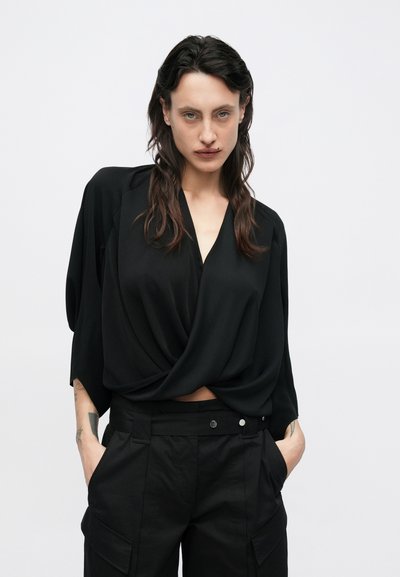 Fekete átkötős blúz mély V-nyakkivágással és redőzött elülső résszel; laza ujjaival és magas derekú fekete cargo nadrággal párosítva.