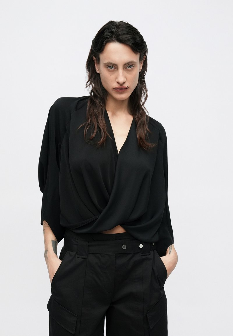 Schwarze Wickelbluse mit tiefem V-Ausschnitt und drapiertem Vorderteil; verfügt über weit geschnittene Ärmel und wird mit hoch taillierter schwarzer Cargo-Hose kombiniert.