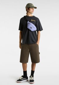 Fialový kostkovaný páskový batoh nošený přes tělo na černém tričku s logem "Vans", spárovaný s hnědými šortkami a černými teniskami Vans.