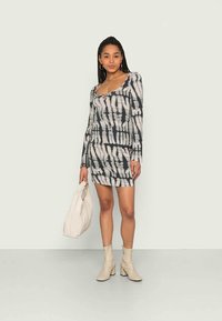 Jovem mulher vestindo um vestido mini de manga longa com estampa tie-dye em preto e branco, segurando uma bolsa branca e usando botas de tornozelo bege, em pé contra um fundo liso.