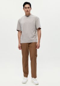 Světle šedé tričko s krátkým rukávem a vysokým výstřihem, spárované s středně hnědými kalhotami. Model v bílých teniskách, stojící na jednobarevném pozadí.