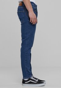 Temno modri jeans s oprijetim krojem, z usnjenim detajlom na pasu in verižnim okrasom, kombinirani z črnimi in belimi športnimi copati.