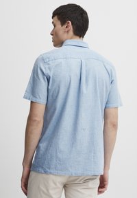 Hele sinine lühikeste varrukatega shirt, mis on valmistatud tekstuuriga kangast, millel on tagaküljel voldi ja klassikaline krae. Pole nähtavaid mustreid ega aktsente.