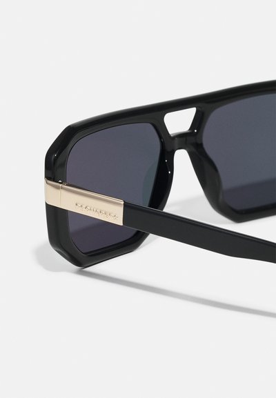 Black sunglasses with an octagonal shape, dark lenses, and a gold-accented temple feature. Glossy finish with a sturdy design.