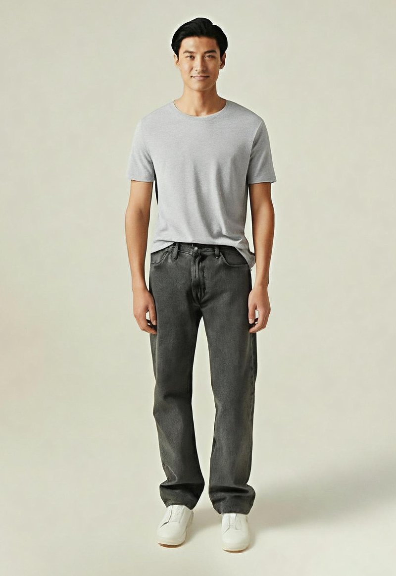 Jeune homme debout, portant un t-shirt gris clair, un jean gris foncé et des baskets blanches sur un fond uni.