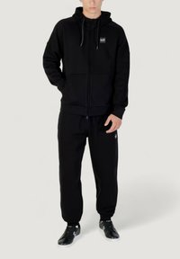 Schwarze Sweatjacke mit Reißverschluss und Kordelzug, kombiniert mit passenden Jogginghosen. Beide Teile haben eine glatte Textur und minimalistische Branding auf der Brusttasche.
