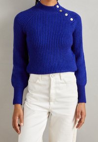 Pull en maille côtelée bleu foncé avec col montant et manches longues. Détail à boutons sur l'épaule. Associé à un denim blanc.