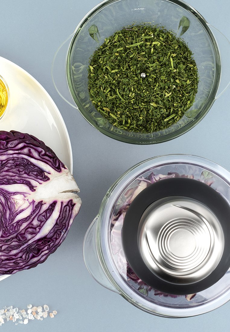 Col morado, cuenco de cristal con hierbas, pequeño cuenco de aceite y cristales de sal sobre un fondo azul claro. Mezcla de texturas y colores presente.