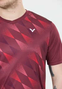 Nærbillede af en person iført en bordeaux sports T-shirt med et rødt geometrisk mønster og et lille hvidt logo på brystet.