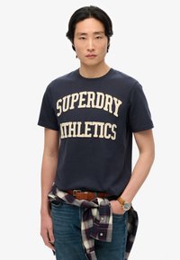 Camiseta de algodón azul marino con "SUPERDRY ATHLETICS" en letras crema, combinada con jeans azules y una camisa de cuadro atada alrededor de la cintura.