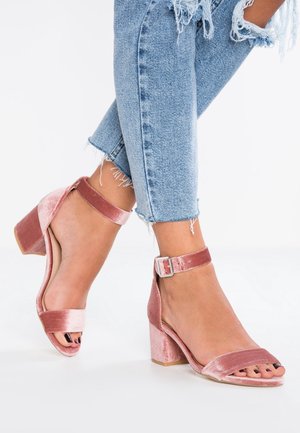 Vaaleanpunaiset samettiset sandaalit, joissa on leveä korko, nilkkaremmi, avoin varvas ja sileä pinta. Varustettu soljella säädettävää istuvuutta varten ja tukevalla korolla.