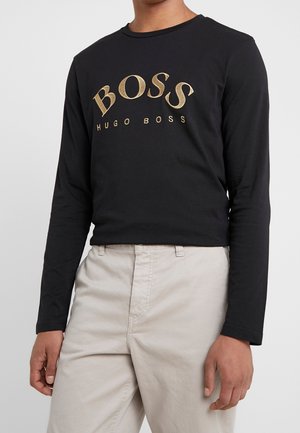 Personne portant une chemise noire à manches longues avec un logo doré « BOSS HUGO BOSS » associée à un pantalon beige clair sur un fond uni.