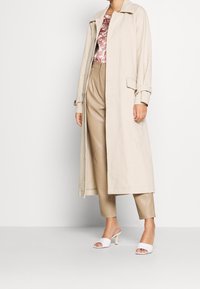 Trench coat lungo beige realizzato in tessuto leggero, abbinato a pantaloni taupe su misura e sandali bianchi con tacco. Presenta un colletto a punte e tasche.