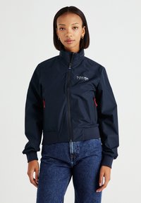 Chaqueta naval resistente al agua con cuello alto, cremallera frontal, puños elásticos y logotipo de la marca. Presenta acentos de cremallera roja y una base ajustada.