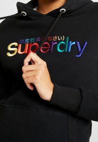Černá mikina s kapucí s vyšívaným vícebarevným logem "Superdry" na hrudi. Má šňůrky na stažení a žebrované manžety. Měkká textura látky.