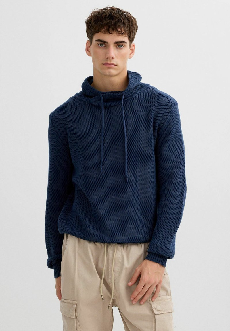 Sudadera con capucha azul marino texturizada con cordón en la capucha y puños acanalados, combinada con pantalones cargo beige claro que tienen bolsillos laterales.