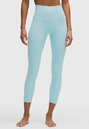 Widok od talii w dół osoby noszącej jasnoniebieskie, krótkie legginsy z wysokim stanem, stojącej boso na jednolitym tle.