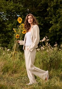 Femme en costume beige et haut blanc tenant des tournesols, debout dans une prairie herbeuse avec des arbres en arrière-plan.