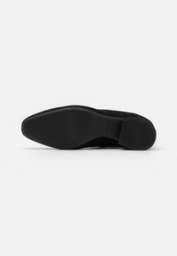 Suela de zapato de ante negro con forma cónica, superficie texturizada y tacón bajo. Cuenta con una base de goma negra lisa y plana para un mejor agarre.