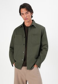 Hombre con chaqueta de botones de color verde oliva sobre un suéter marrón oscuro y pantalones beiges, de pie con las manos en los bolsillos frente a un fondo blanco.