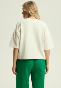Kremowa, skrócona koszulka z krótkim rękawem, charakteryzująca się luźnym krojem i teksturowanym materiałem. Noszona z zielonymi spodniami, z prostymi detalami designu.