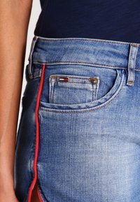 Lähikuva sinisistä denimfarkuista, joissa on etutasku, pieni logo, punainen sivusauma ja henkilö, jolla on tumma yläosa.