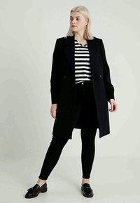 Svarte lange frakk med skreddersydd design, med notched krage og dobbelte knapper, båret over en stripete topp og svarte leggings.