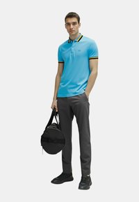 Polo shirt azzurro con strisce gialle e nere in contrasto sul colletto, abbinato a pantaloni grigi scuri e una borsa rotonda nera.