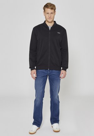 Černá bunda na zip s vysokým límcem, kombinovaná s modrými džínami a bílými teniskami, s elegantním designem a ležérním střihem.