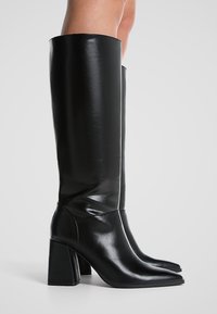 Bottes noires montantes en cuir lisse avec une pointe effilée et un talon carré épais. Présente un design épuré et une finition brillante.