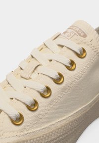 Κοντινή λήψη μπεζ καμβά αθλητικού παπουτσιού με λευκά κορδόνια, χρυσές τρύπες, υφή στη σόλα από καουτσούκ και ορατή σήμανση "Converse" στη γλώσσα.