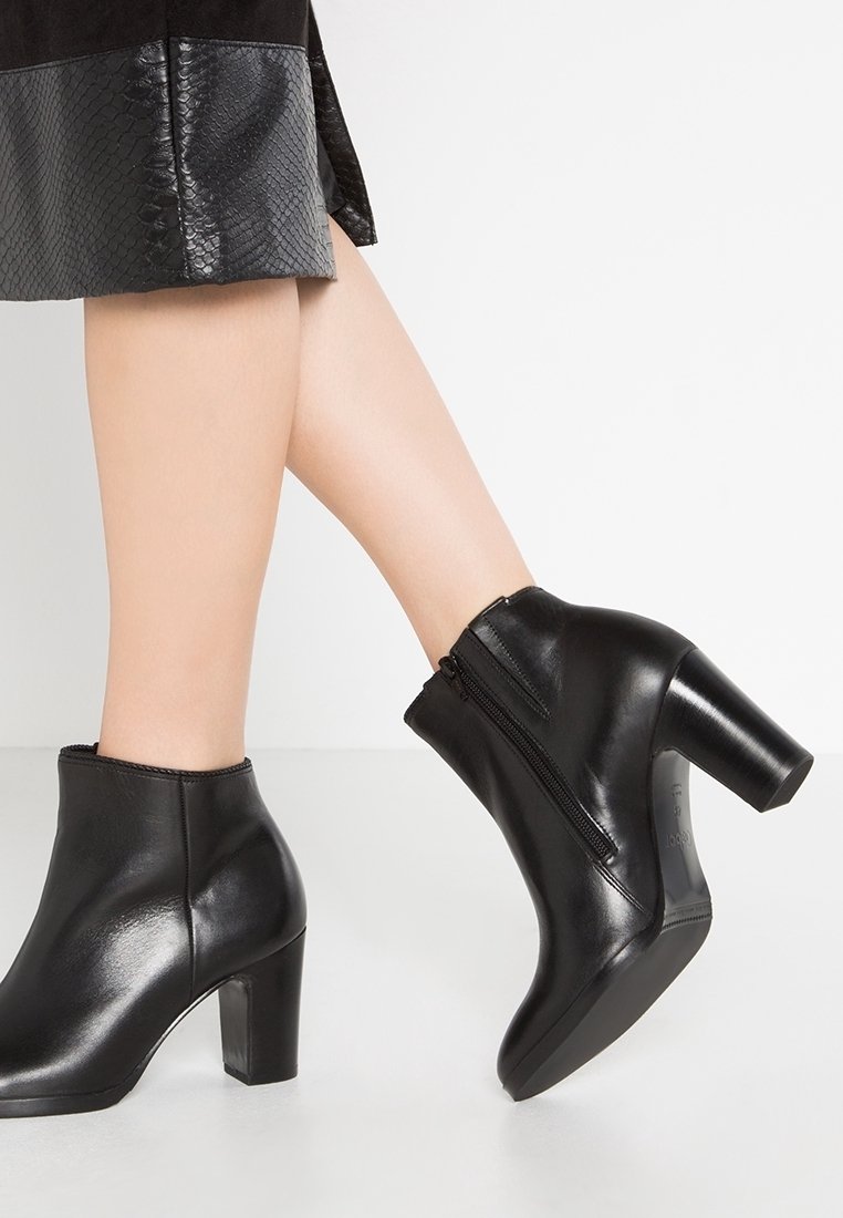 Jambes portant des bottines en cuir noir avec des talons carrés et des fermetures éclair latérales, associées à une jupe texturée noire.