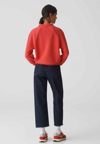 Rode textuurtrui met een hoge kraag, gecombineerd met donkere spijkerbroek en multicolore sneakers. De outfit heeft een ontspannen pasvorm en een casual stijl.