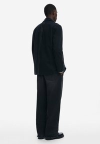 Chaqueta de mezclilla negra con corte recto, costuras visibles y pantalones negros a juego. Tela oscura y texturizada con dobladillos ligeramente deshilachados y bolsillos.