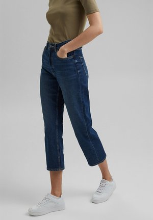 Frau trägt kurze blaue Jeans, olivgrünes kurzärmliges Oberteil und weiße Sneakers, steht mit einer Hand in der Tasche vor grauem Hintergrund.