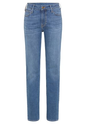 Persona de pie con los brazos cruzados, vestida con una sudadera rosa pálido, vaqueros azules y zapatillas blancas sobre un fondo gris liso.