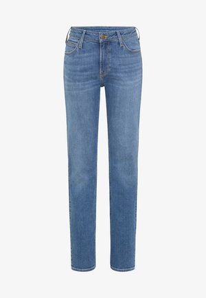 Persona de pie con los brazos cruzados, vestida con una sudadera rosa pálido, vaqueros azules y zapatillas blancas sobre un fondo gris liso.
